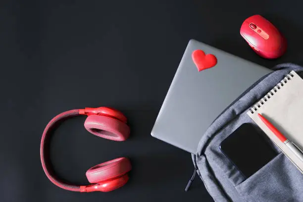 Photo of Gray trendy laptop backpack with a mobile phone, a notepad, a pen and a laptop on a black background next to red headphones and a mouse. Soft laptop sticker in the shape of a heart.