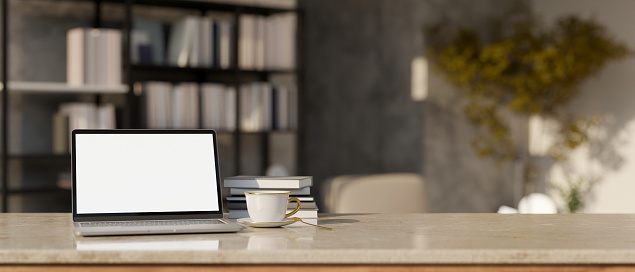 Workspace tabletop with laptop white screen mockup, coffee cup, books and copy space over blurred modern living room with bookshelves in the background. 3d render, 3d illustration