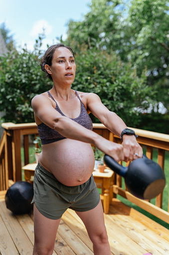 An athletic multiracial pregnant woman does kettlebell swings during an at home strength and cardio workout outside on the back patio of her home.