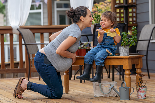 A pregnant multiracial woman and her toddler daughter giggle while gardening and caring for potted plants together on the back patio of their home.