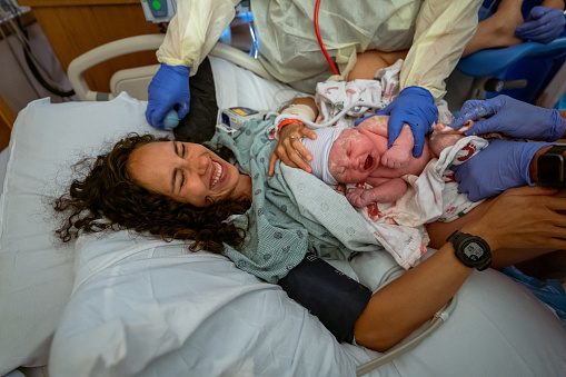 A beautiful multiracial woman of Hawaiian descent smiles and cries with gratitude as her medical team places her healthy newborn baby on her chest for skin to skin bonding directly after delivery.