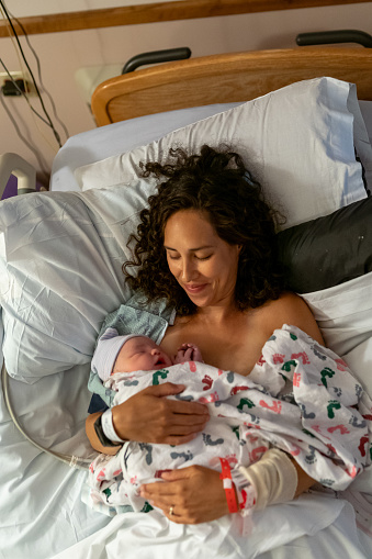 A beautiful multiracial woman lying in a hospital bed smiles affectionately at her newborn baby who is sleeping peacefully in her comforting embrace as they enjoy bonding with skin to skin contact.