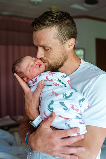A proud new father stands in a hospital room and lifts his sleeping newborn baby close to his face so he can give the child a kiss on the cheek. The family is preparing for discharge and is excited to bring the one day old baby home.
