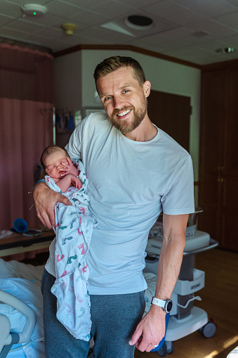 Portrait of a happy new dad smiling directly at the camera while holding his newborn baby in one arm. The family is preparing for discharge from the hospital and are excited to go home.