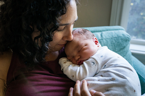 Cropped shot of a multiracial woman of Pacific Islander descent holding her Eurasian newborn baby against her chest and lovingly looking at the sleeping child.