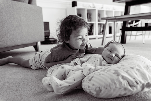 A cute Eurasian toddler girl lies on the floor in the living room at home and lovingly interacts with her baby brother. Image is black and white.