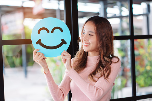 Asian woman holding blue happy smile face on paper cut, user giving good feedback rating, think positive , customer review, assessment, of mental health day, Compliment Day, satisfaction concept.