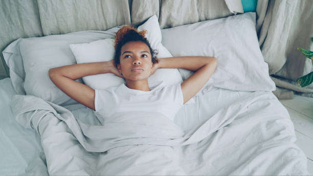 istock Sad African American lady is lying in bed and sighing looking at ceiling. Comfortable bed, pillows and sheets are visible. 1438289538