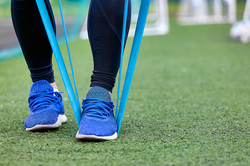 Close-up leg athlete performing an exercise with sports elastic band on green artificial surface of stadium. Training process before the competitions of the athlete football player in the fresh air