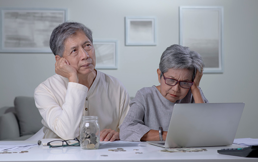 Worried Asian senior couple reading financial documents or notification letter with bad news from bank while paying bills online on laptop, retired husband and wife having money problems