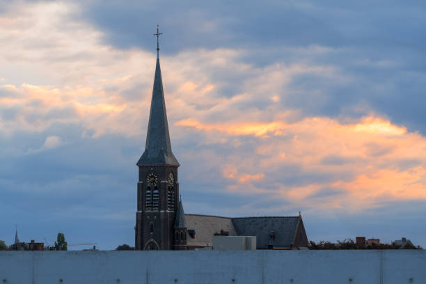 A white baroque-style church tower with a dark, ornate spire rises above green treetops under a dramatic sky filled with thick clouds. The clock on the tower and the classic architecture convey a timeless, tranquil atmosphere typical of European villages.