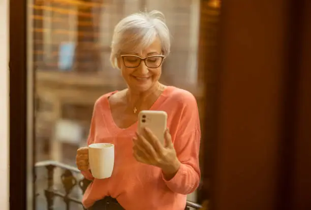happy senior Caucasian woman in apartment on balcony, drinking coffee while checking her cell phone