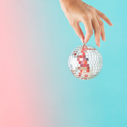 Minimal abstract Xmas card with female hand holding a disco ball on an isolated iridescent turquoise-pink background. New Year's Eve holiday entertainment concept. Christmas decoration.