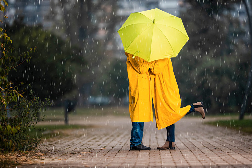 Couple in yellow raincoats kissing under the umbrella during rainy day at the park. Copy space.