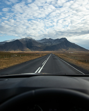 A vertical shot of a car on the road surrounded by rocky hills under a blue cloudy sky in Iceland