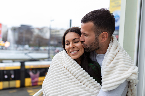 Romantic couple on a winter holiday. Man and woman standing together in a hotel room balcony wrapped in blanket. Couple embracing and smiling.