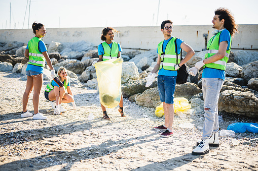 People cleaning up the beach, volunteers collecting the waste on the coast line, young generation z friends working in team aware of the pollution produced by the plastic industry.