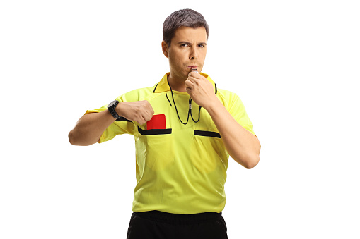 The football referee in his referee shirt with whistle, moves the football to the area of game play on a very green football field shown in the background.  Room for copy space.