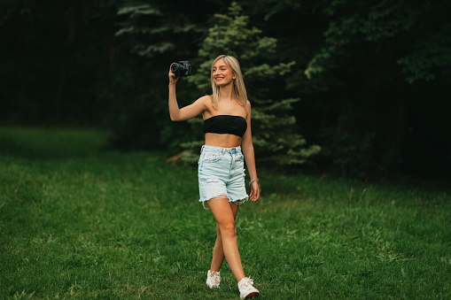 Smiling young casually dressed woman taking photos with a digital camera and enjoying a sunny summer day in nature