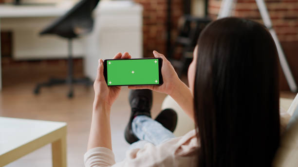 istock Woman sitting on sofa while holding smartphone with green screen mockup template background 1438337500