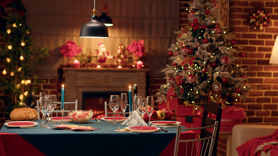 Empty traditional Christmas dinner table inside decorated living room with holiday garlands and dinnerware. Interior of traditional and authentic season cozy setting celebrating religious event.