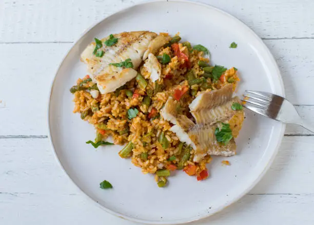 Pan fried natural fish fillet with brown rice and mixed vegetables on a white plate isolated on white wooden background from above. Closeup with copy space