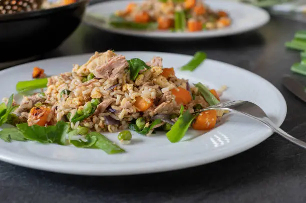 Delicious homemade healthy rice dish. Fried brown rice with egg, vegetables and tuna. Served ready to eat on a plate on kitchen table. Front view with blurred background