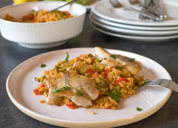Healthy homemade fish meal with pan fried, natural fish fillet and vegetable rice. Cooked with brown rice, tomatoes, bell peppers, onions, garlic, herbs and green beans. Served ready to eat on a plate on kitchen table