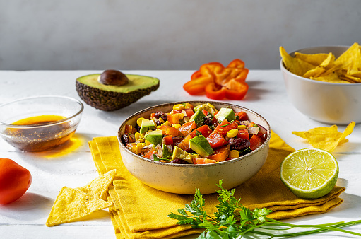 Cowboy caviar is traditional Mexican vegetable salad in bowl made with avocado, black beans, bell pepper, corn, coriander, lime juice and dip. White wooden background, yellow napkin
