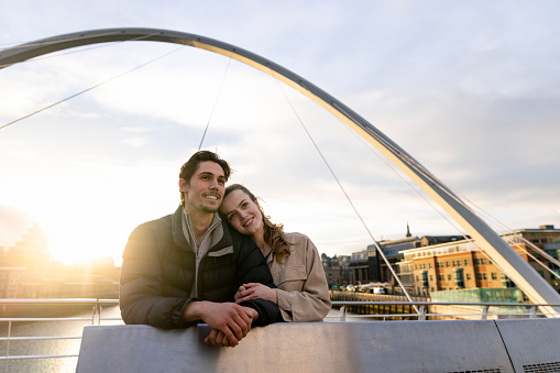 A wide-view shot of a young couple standing side by side looking at the view together on a bridge in the city, they are in love.