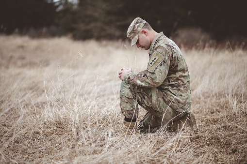 A shallow focus shot of a young soldier praying while kneeling on a dry grass