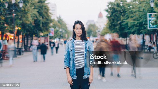 istock portrait of tired woman student standing alone in city center and looking at camera with straight face while crowds of men and women are whizzing around. 1438350179