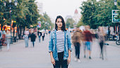 portrait of tired woman student standing alone in city center and looking at camera with straight face while crowds of men and women are whizzing around.