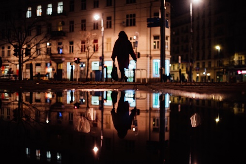 A silhouette of a woman heading home with grocery bag and her reflection on a puddle at night