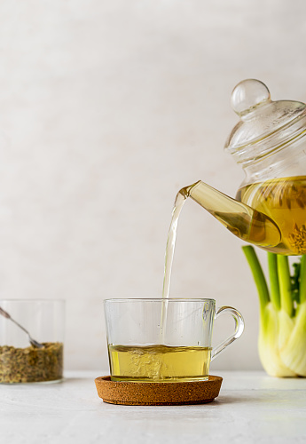 Fennel tea in a glass cup, fresh fennel bulb, seeds and teapot pouring tea to cup. White wooden table with light background. Vertical banner. Copy space