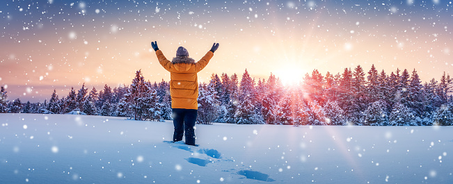 A woman stands on a snowy field with her hands raised up. Panoramic background.
