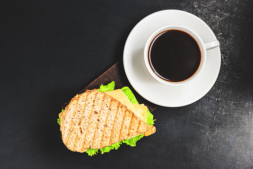 Grilled sandwich with a cheese, salad lettuce, ham and coffee cup on dark background, top view. Lunch concept with copy-space.