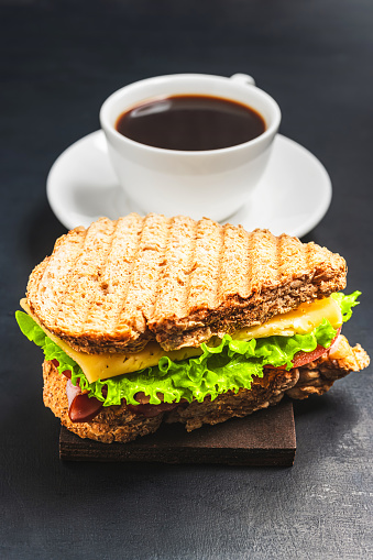 Grilled sandwich with a cheese, salad lettuce, ham and coffee cup on dark background. Lunch concept with copy-space.
