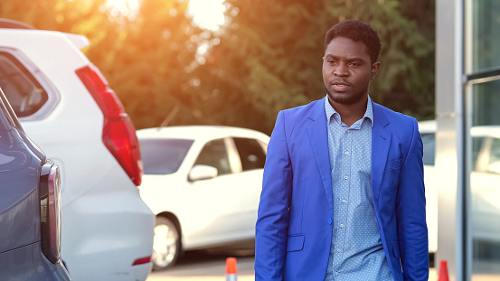 African American male customer searches new nice automobile on luxury salon open ground. Black man with serious expression examines cars on parking lot, sunlight