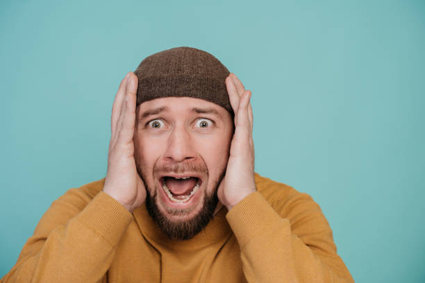 istock Shocked young man with opened mouth in sweater and hat screaming with horror face expression standing against turquoise studio backdrop. Isolated close up shoot of scared male disappointed by bad news 1438371191