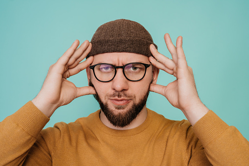 Close up portrait of thoughtful young man in winter hat, glasses and sweater touching temples by fingers concentrated on tasks, isolated over turquoise background. Creative people, mindfulness.