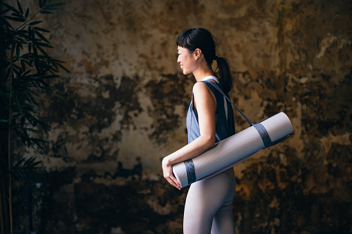 Smiling Asian woman in sportswear holding exercise mat while walking at yoga studio.
