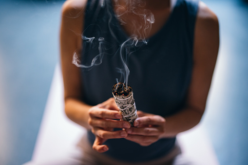 From above photo of unrecognizable Asian woman preparing for meditating on a exercise mat while holding white sage incense at yoga studio.