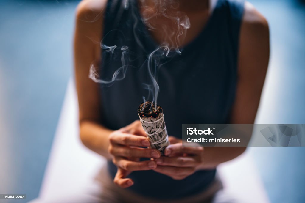 Close Up Photo Of Woman Hands Holding Lighted Bundle Of Sage Before Yoga Session From above photo of unrecognizable Asian woman preparing for meditating on a exercise mat while holding white sage incense at yoga studio. Sage Stock Photo