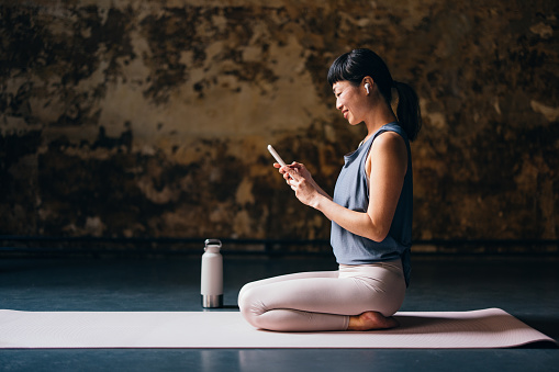 Side view of happy Asian woman woman in sportswear sitting on a exercise mat and listening to music on her smartphone before yoga session.