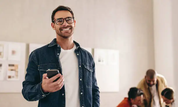 Smiling businessman with eyeglasses holding a smartphone while standing in an office. Happy young businessman working in a creative and modern workplace with his colleagues.