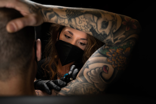 Concentrated tattoo artist with facial mask tattooing a man in a dark studio
