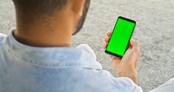 Close-up of man looking at smartphone Over the shoulder view of man looking at green screen of smartphone while sitting in park. man looking at phone over shoulder stock pictures, royalty-free photos & images