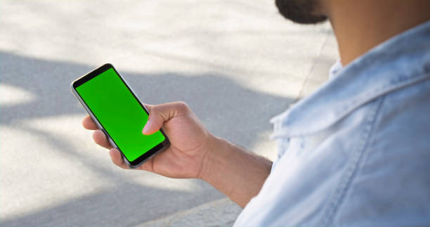 Close-up of man looking at smartphone Over the shoulder view of man looking at green screen of smartphone while standing in park. man looking at phone over shoulder stock pictures, royalty-free photos & images