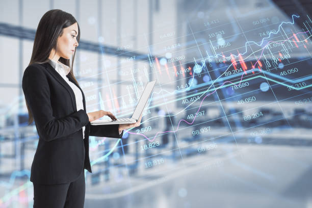 Attractive thoughtful young european businesswoman with laptop computer and creative candlestick forex chart index hologram on blurry office interior background. Trade, business market and finance analysis concept. Double exposure. stock photo